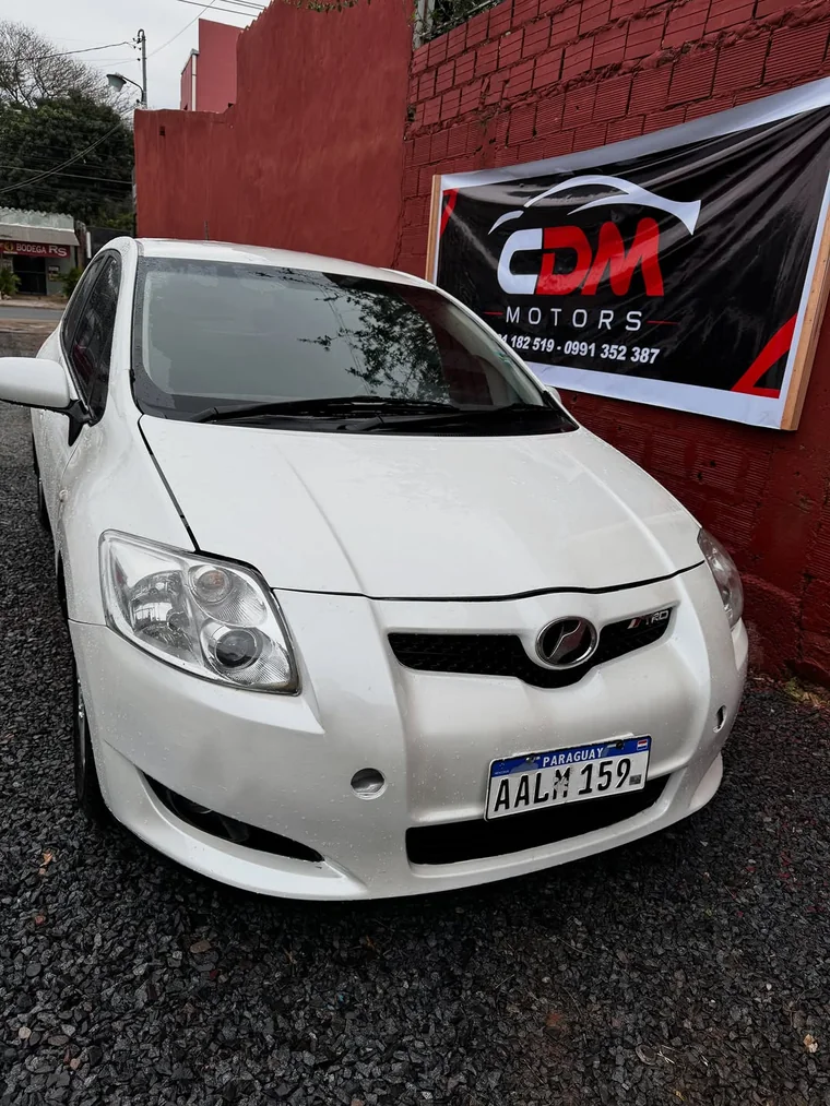 Toyota Auris blanco estacionado en el lote de CDM Motors, frente al cartel con los dos teléfonos